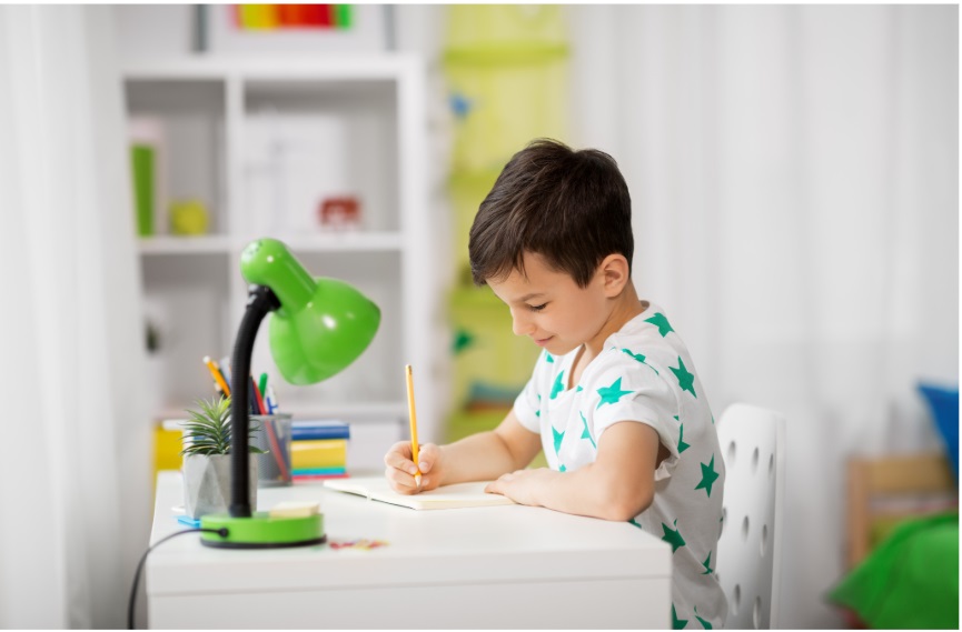 A young boy drawing with a pencil at a white desk with a green lamp in a bright study room